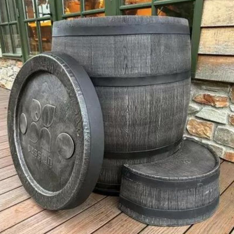 "The Ice Man's Barrel by Passion Ice Baths and Wim Hof, showcasing a large, empty barrel-shaped ice bath with a detachable lid, set on a wooden deck with a stone wall and windows in the background. The barrel has a rustic wood finish and is designed for cold water immersion therapy."