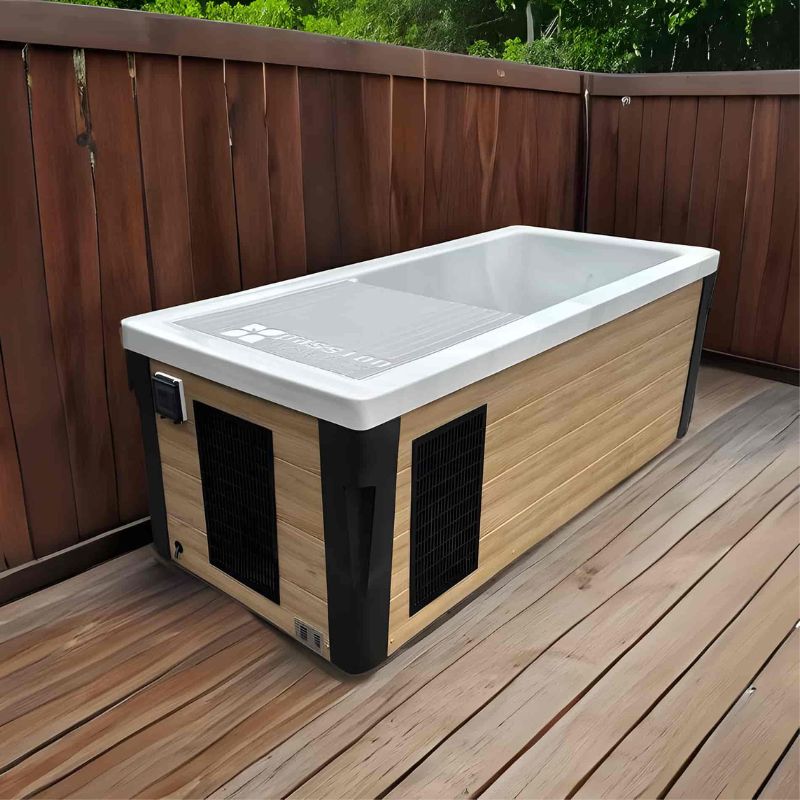 "The Breeze Ice Bath with current by Passion Ice Baths and Wim Hof, featuring a rectangular design with a wood-look exterior and white interior, placed on a wooden deck surrounded by a wooden fence."