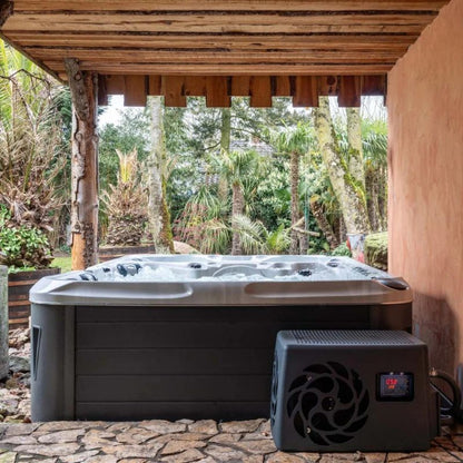 "The Elevate Contrast Ice Bath with current by Passion Ice Baths and Wim Hof, set outdoors under a wooden canopy. The dual-zone bath, partially visible, features bubbling water. The chiller unit is placed beside the bath, with a digital temperature display. The background shows lush greenery and trees."