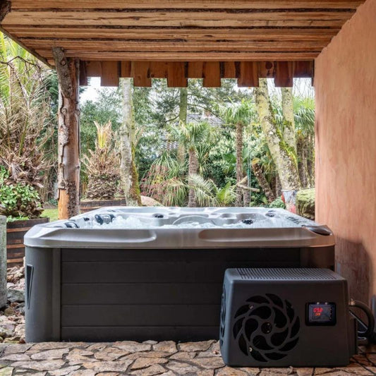 "The Elevate Contrast Ice Bath with current by Passion Ice Baths and Wim Hof, set outdoors under a wooden canopy. The dual-zone bath, partially visible, features bubbling water. The chiller unit is placed beside the bath, with a digital temperature display. The background shows lush greenery and trees."