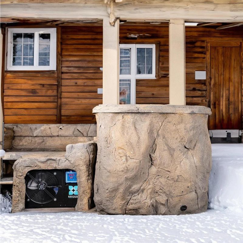 "The Faith - Rock Ice Bath and Chiller by Passion Ice Baths and Wim Hof, positioned outside a wooden cabin in a snowy setting. The ice bath features a rock-textured design with an integrated chiller unit, blending seamlessly with the rustic cabin environment."