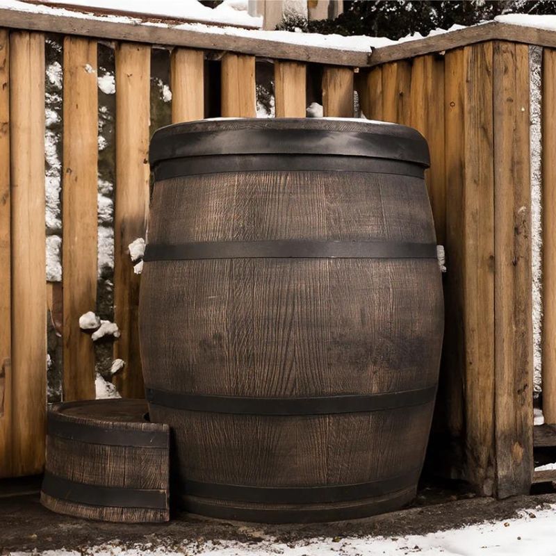 "The Ice Man's Barrel by Passion Ice Baths and Wim Hof, a rustic wood-finished ice bath, set outdoors by a wooden fence with snow on the ground and railing. A detached, matching wooden step is placed beside the barrel."