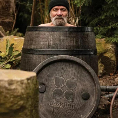 "Wim Hof, also known as The Ice Man, sitting in The Ice Man's Barrel by Passion Ice Baths. The barrel-shaped ice bath, made of rustic wood, is set in an outdoor natural setting with plants and rocks. The detachable lid is placed in front of the barrel, displaying the Passion Ice Baths logo."