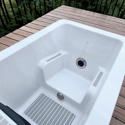 "Close-up of the interior of the Breeze Ice Bath with current by Passion Ice Baths and Wim Hof, showing the white, contoured seating area, water jets, and drain, set on a wooden deck with greenery in the background."